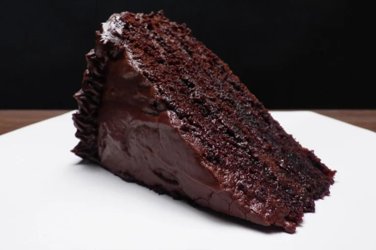 A close-up photo of a sliced decadent chocolate cake. The cake is dark and moist with a rich chocolate frosting. A single raspberry sits atop the slice.